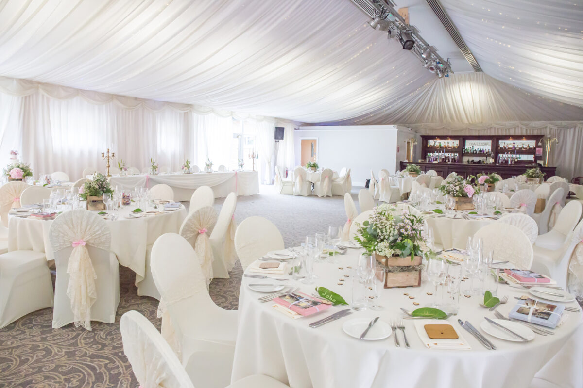The Villa Wrea Green in Lancashire offers a spacious, elegantly decorated wedding reception hall with round tables draped in white linens, adorned with floral centerpieces. The ceiling is covered with white drapes, and a bar area is visible in the background. Soft lighting creates a romantic ambiance. Image by Andy Wade Photography.