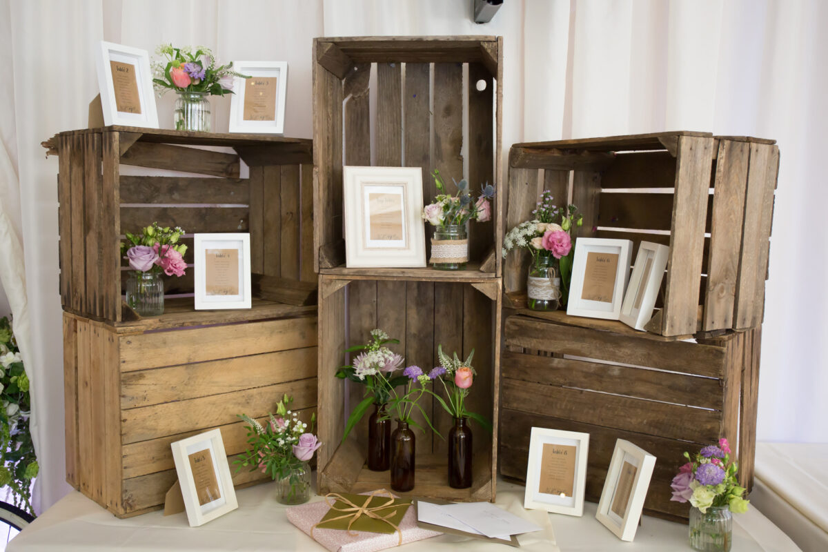 A display of wooden crates arranged on top of each other forms shelves, holding various framed signs, small floral arrangements in vases, and rustic decorative items. The setting appears to be inside a tent with white drapery in the background, reminiscent of an event at The Villa Wrea Green in Lancashire. Image by Andy Wade Photography.