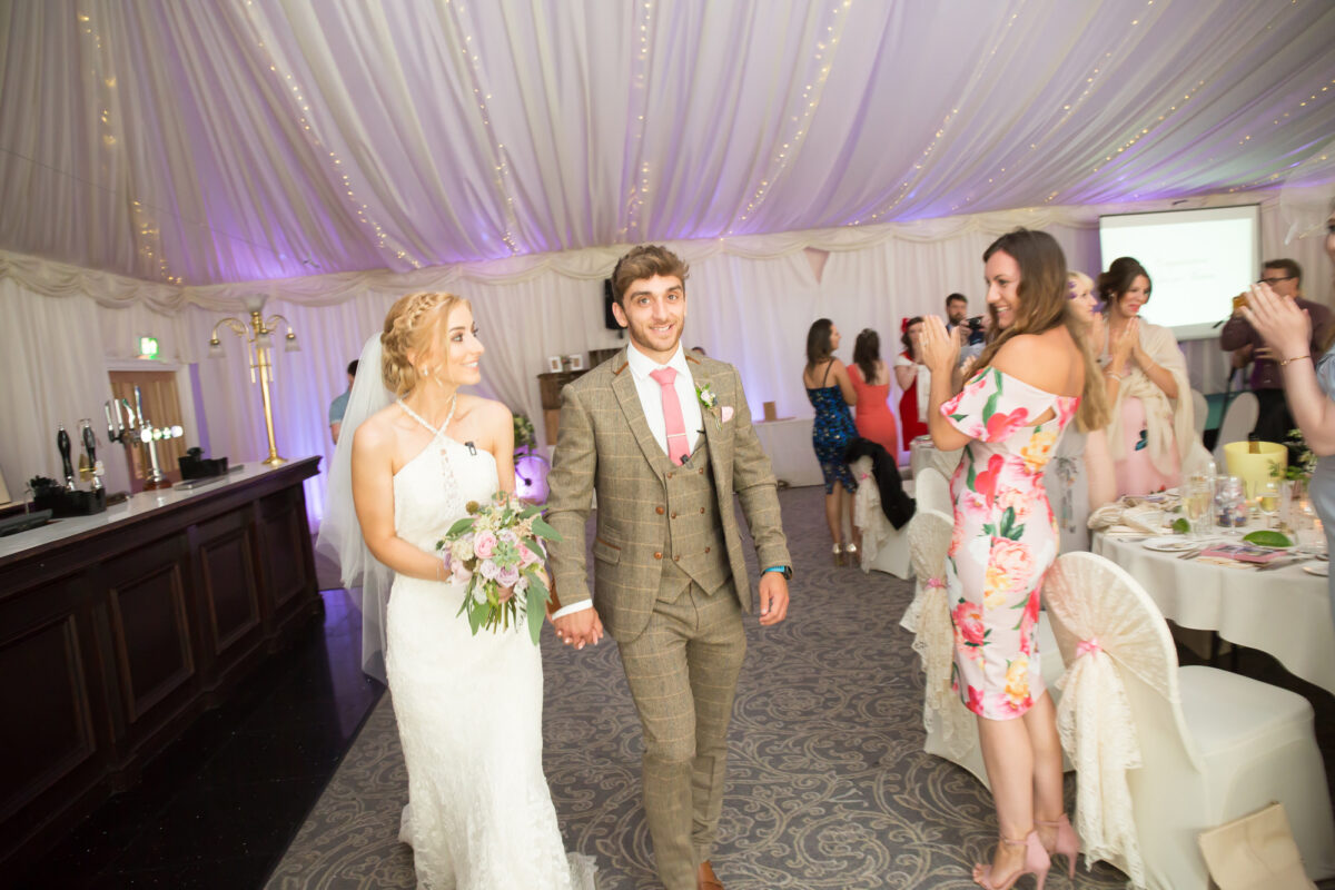 A bride in a white dress and groom in a brown checked suit with a pink tie are holding hands and smiling as they walk through a decorated wedding reception tent at The Villa Wrea Green, Lancashire. Guests are clapping in the background, with fairy lights and elegant drapery setting the scene. Image by Andy Wade Photography.