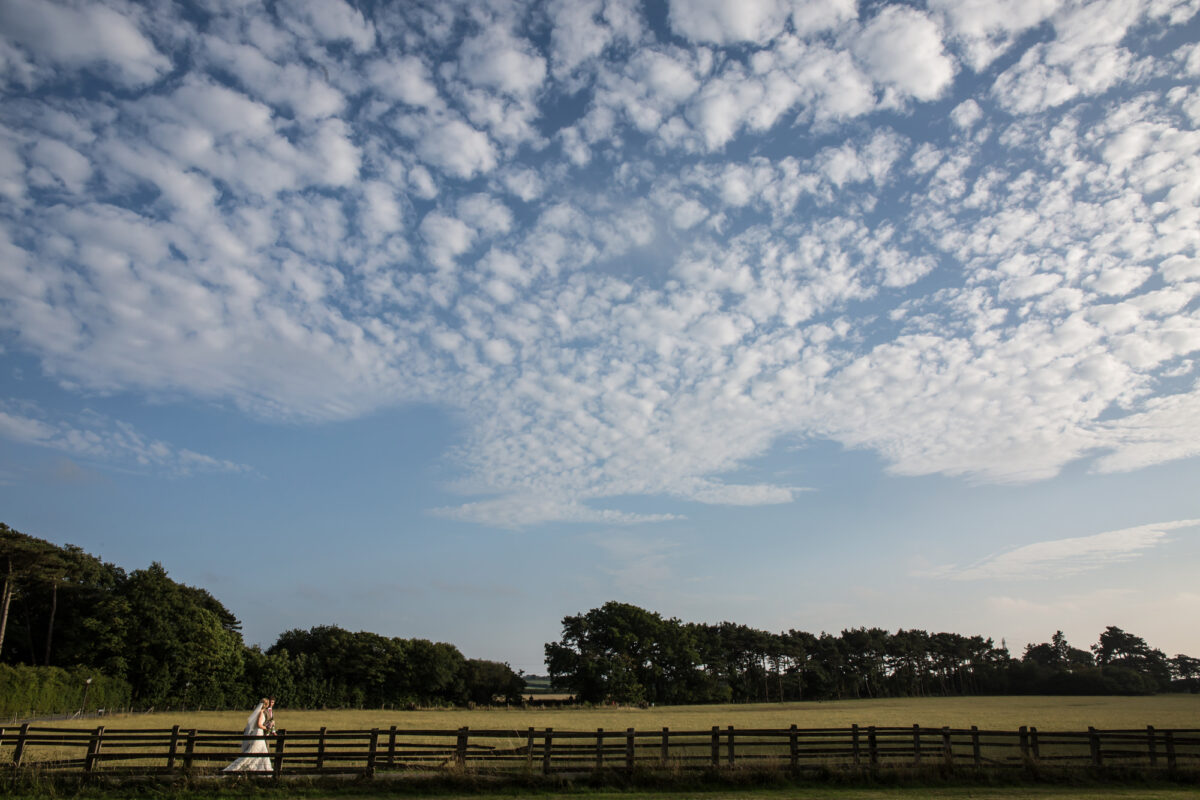 A solitary bride in a white dress walks along a wooden fence in an expansive, open field under a sky filled with scattered, fluffy clouds. At the edge of the field, trees line the distance, creating a serene and calm scene reminiscent of the picturesque surroundings of The Villa Wrea Green in Lancashire. Image by Andy Wade Photography.