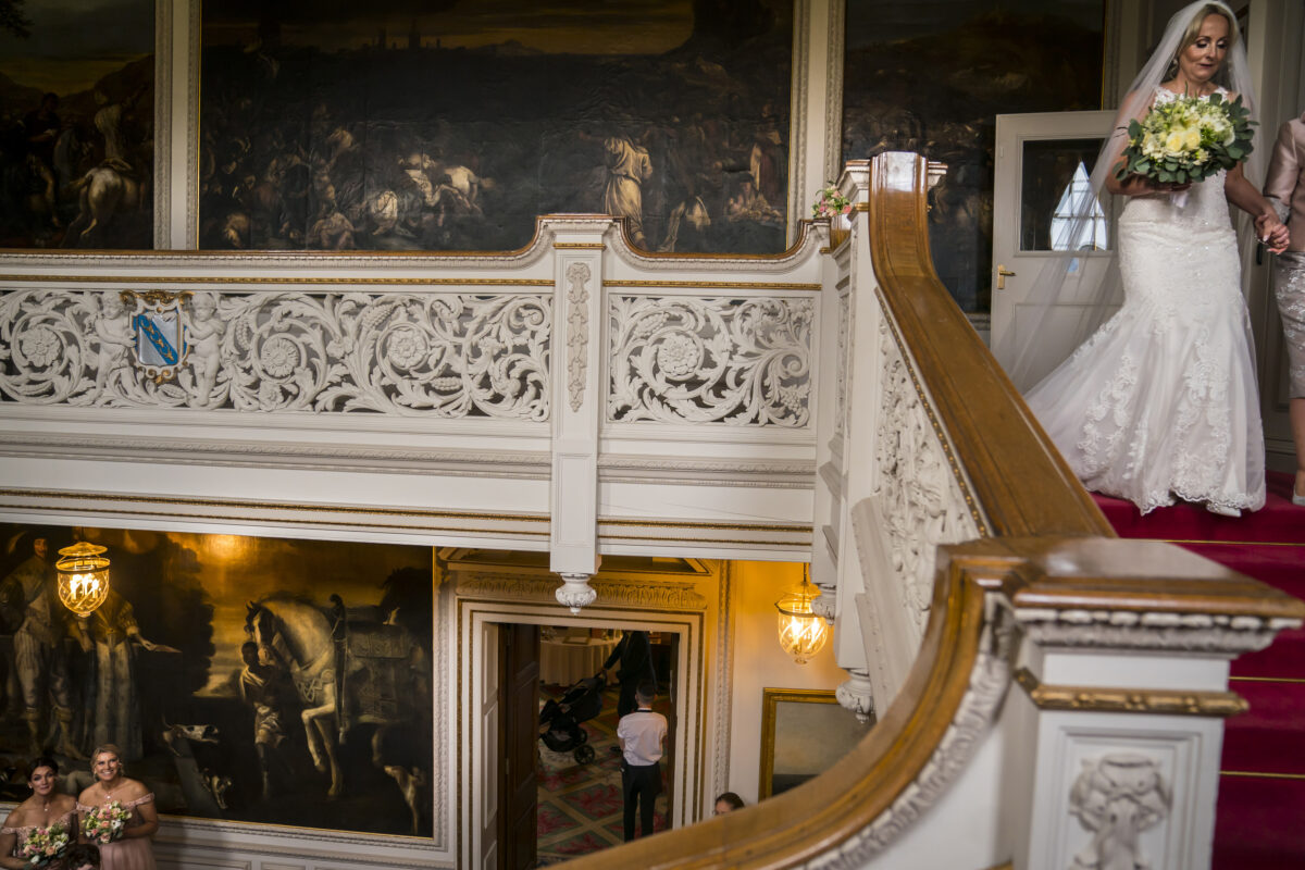 A bride in a white dress and veil descends an ornate staircase with intricate carvings and red carpeting in the grand, historic Knowsley Hall, Lancashire. Below, bridesmaids in light dresses wait, holding bouquets, with a lit chandelier and doorway in view amidst large paintings on the walls. Image by Andy Wade Photography.
