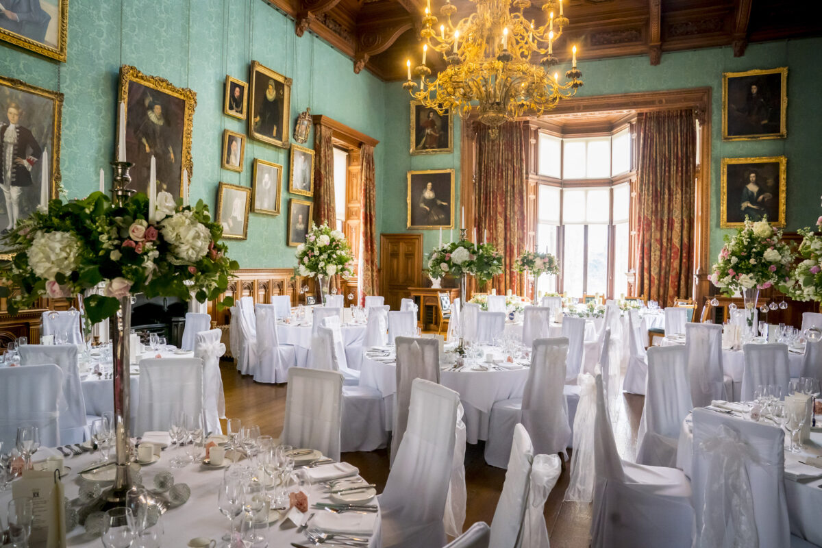 A grand banquet hall at Knowsley Hall with tall windows letting in natural light, adorned with elegant chandeliers and classical paintings on teal walls. Tables are set with white cloths, floral arrangements, and white-draped chairs, ready for a formal event or wedding reception in Lancashire. Image by Andy Wade Photography.