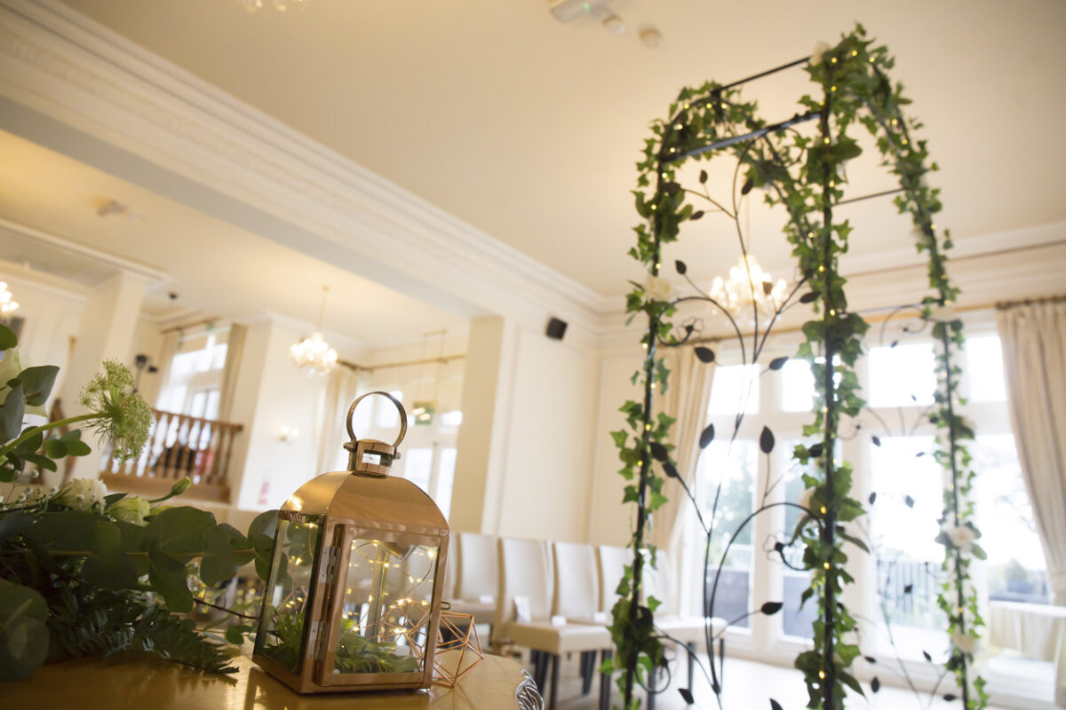 A bright and airy room in the West Tower, Lancashire, decorated for a wedding ceremony. A metal arch adorned with green ivy and leaves stands prominently. A lantern with a lit candle sits on a nearby table. Rows of white chairs are aligned, and large windows in the background let in natural light. Image by Andy Wade Photography.