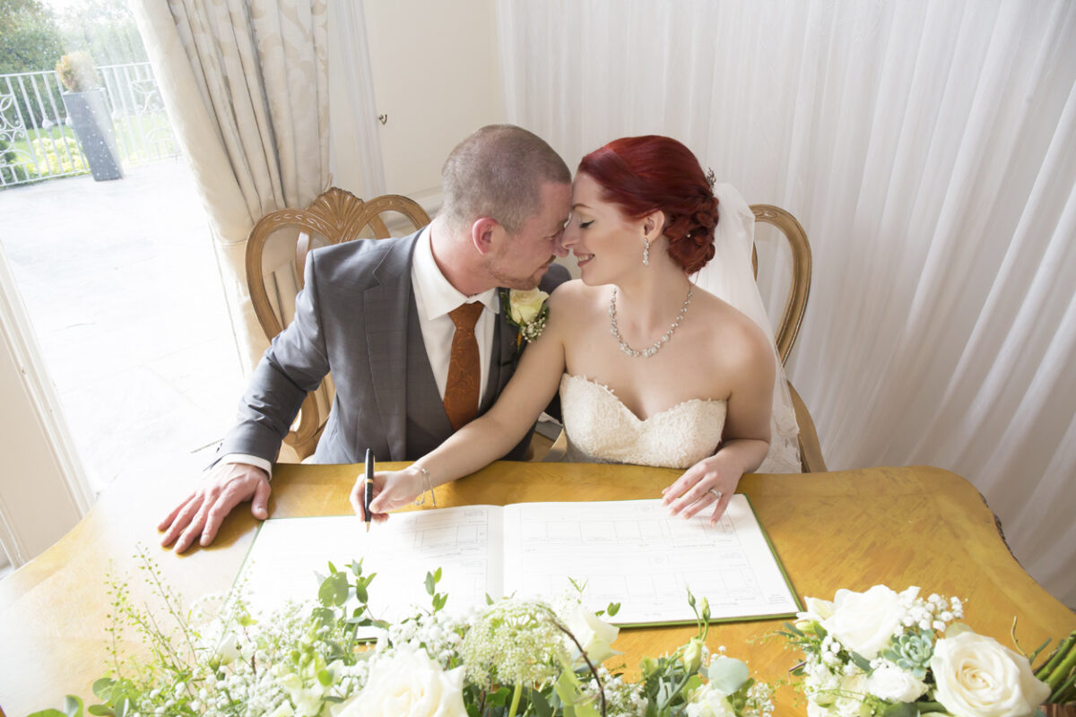 A bride and groom sit at a table in Lancashire's West Tower to sign their marriage certificate. The groom, wearing a grey suit, leans in to affectionately touch heads with the bride, who is in a strapless white gown. The table is decorated with white flowers, and soft light filters through sheer curtains. Image by Andy Wade Photography.