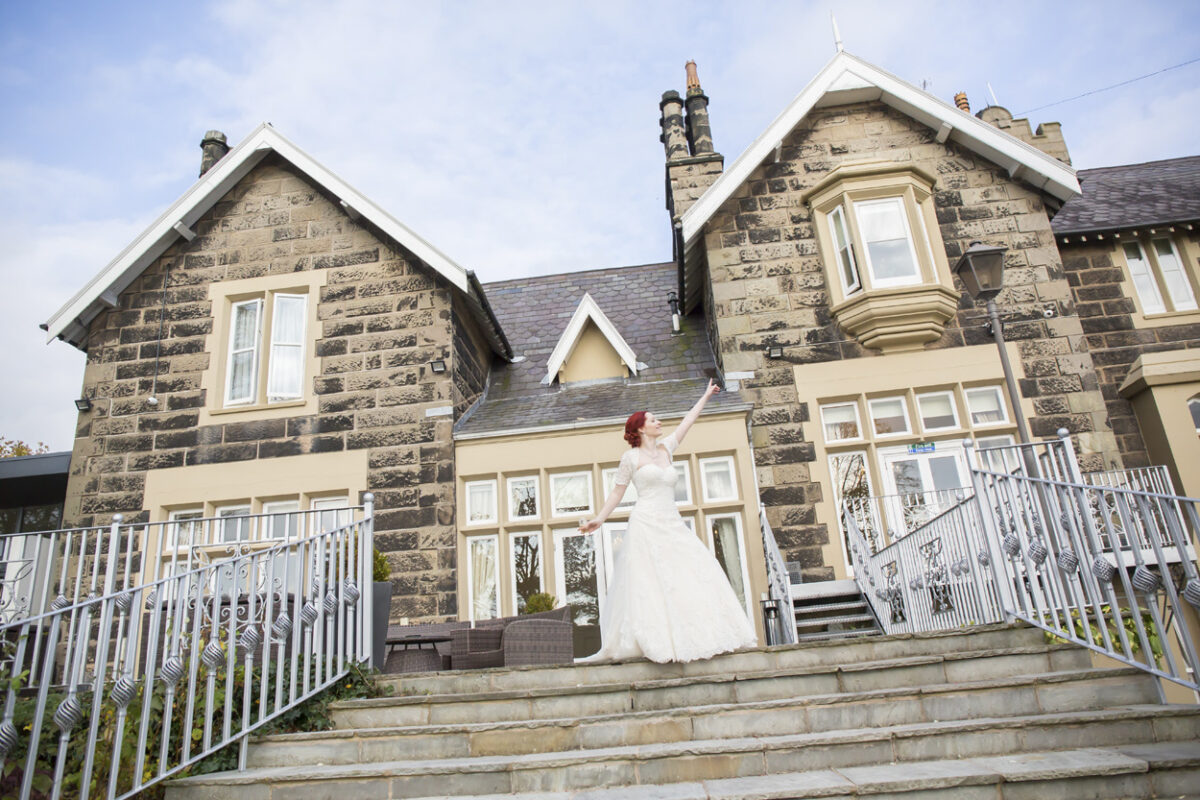 A bride stands on the steps of a large stone house with beige and white trim at West Tower in Lancashire, raising one arm as she faces forward in her wedding dress. The house has multiple windows, dormers, and chimneys, set against a partly cloudy sky. Image by Andy Wade Photography.