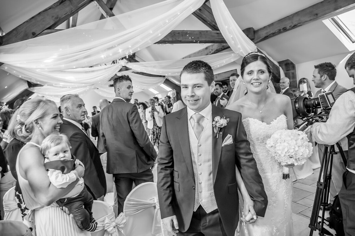 A smiling bride and groom walk down the aisle of Beeston Manor, holding hands. Guests cheer in the background, a woman holding a baby sits on the left. The venue features exposed wooden beams and draped fabric with lights. The mood is celebratory and joyful in this charming Lancashire setting. Image by Andy Wade Photography.