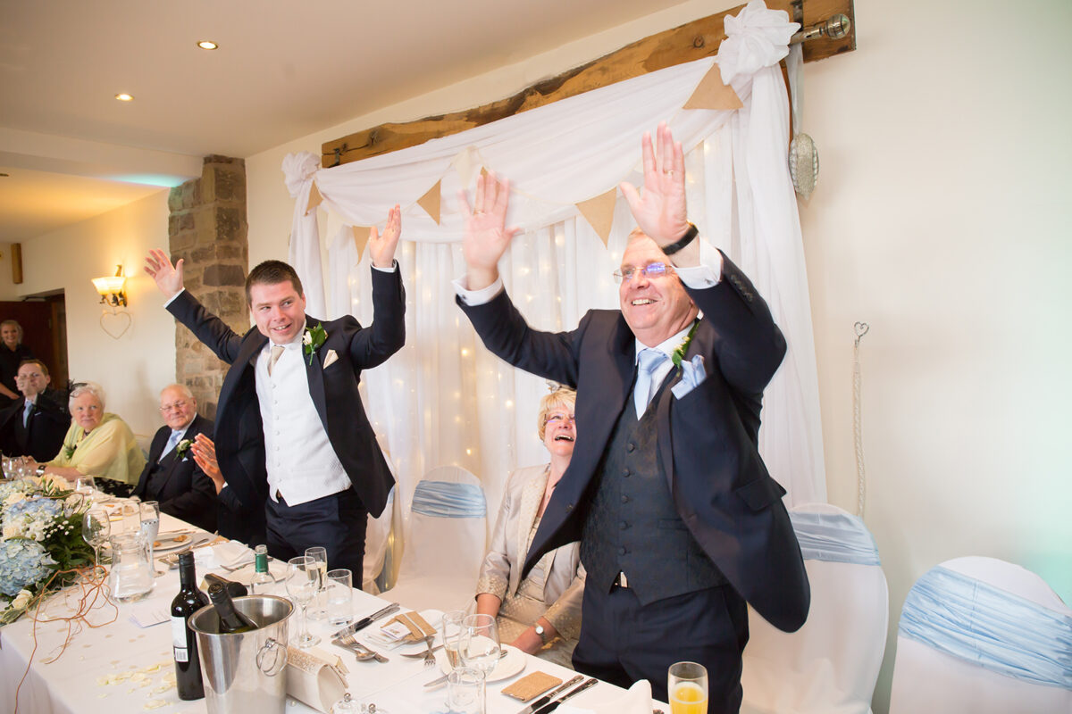 Two men in formal suits stand at a wedding reception table at Beeston Manor, raising their arms and smiling joyfully. Guests seated nearby, including an elderly woman, look on with smiles. The decorated backdrop with draped fabric and fairy lights adds a festive atmosphere to the Lancashire scene. Image by Andy Wade Photography.