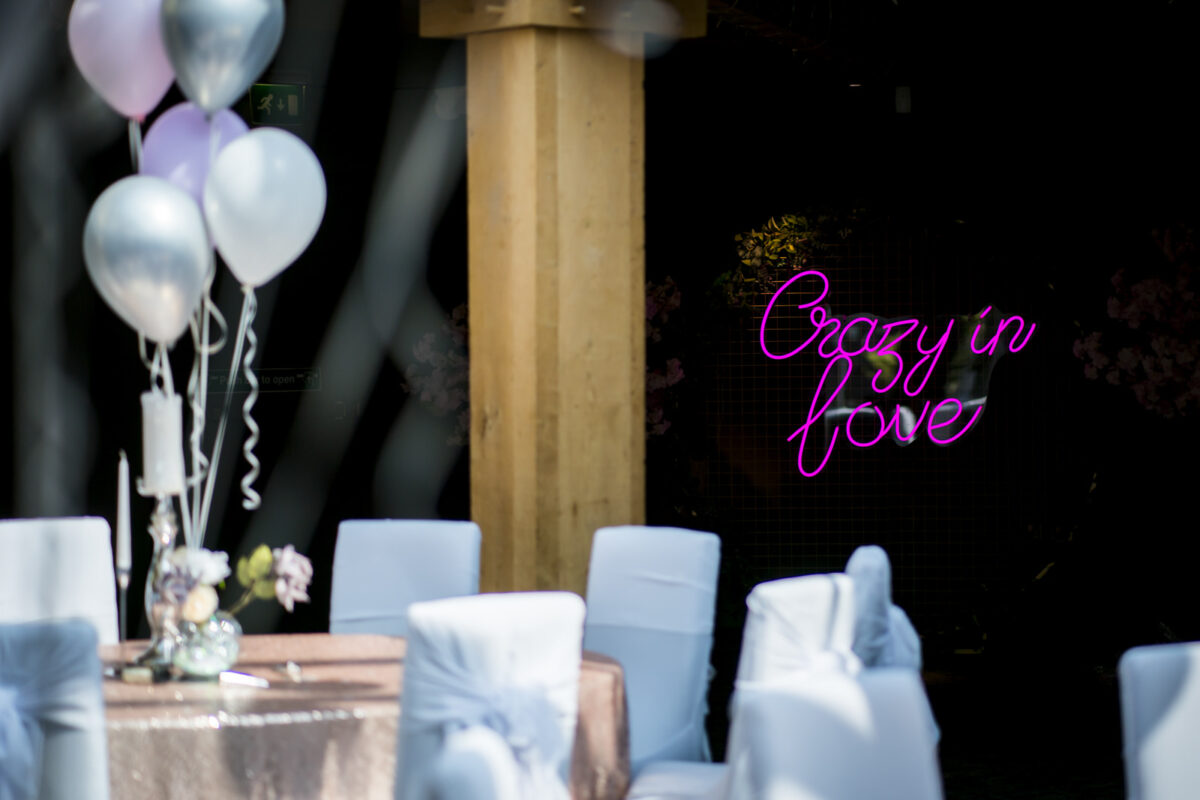 A decorated outdoor space at The White Hart in Lancashire features white satin-covered chairs and a table draped with a sequin tablecloth, adorned with a vase of flowers. Silver and white balloons are tied together on the side, while in the background, a pink neon sign reads "Crazy in Love. Image by Andy Wade Photography.