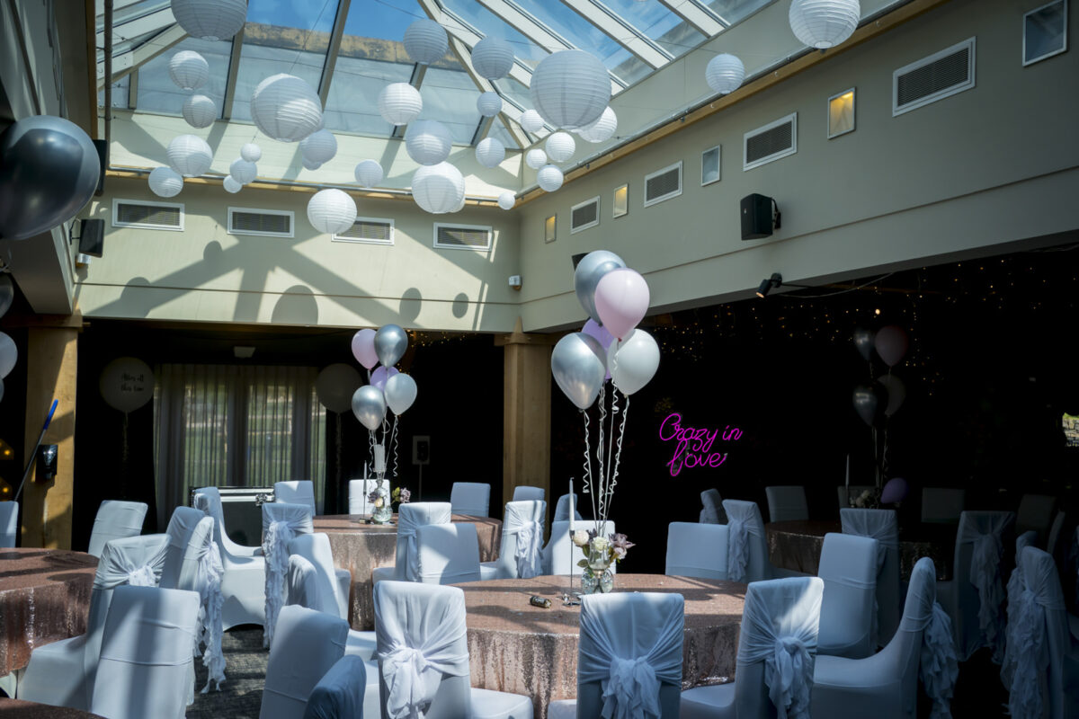 A decorated indoor event space at The White Hart, Lancashire, features round tables covered in pink and silver tablecloths with chairs adorned with silver bows. White and pink balloons float above the tables, while white paper lanterns hang from the glass ceiling. A neon sign reading "Crazy in Love" glows in the background. Image by Andy Wade Photography.