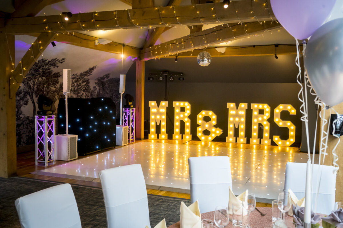A decorated wedding venue at The White Hart in Lancashire features large illuminated "MR & MRS" letters on a dance floor. The ceiling is adorned with string lights, and the seating area has white covered chairs and neatly arranged tables with place settings. Balloons are also visible. Image by Andy Wade Photography.