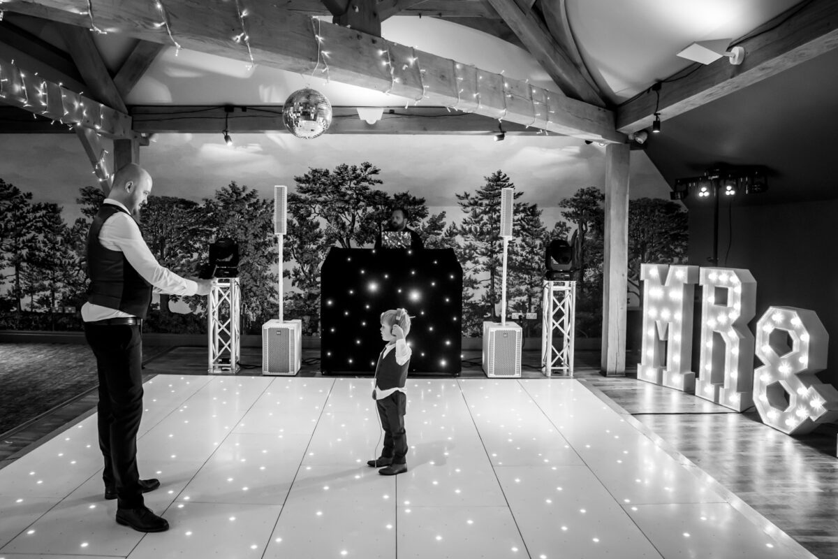 A black and white photo shows a man and a young child on a lighted dance floor beneath a disco ball at The White Hart. A DJ booth with speakers is in the background, alongside illuminated letters "MR &". The room, located in Lancashire, features exposed wooden beams and fairy lights strung overhead. Image by Andy Wade Photography.