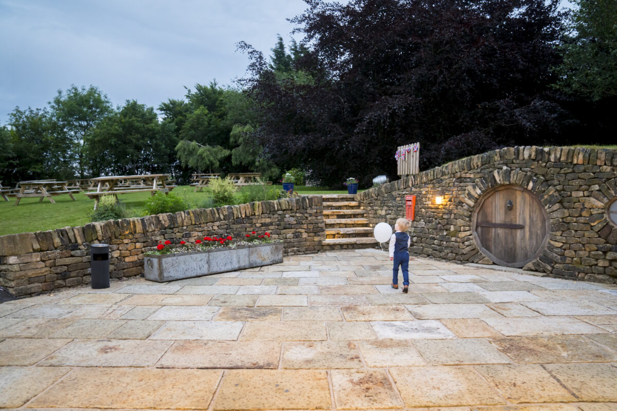 A young child holding two white balloons stands in a courtyard with stone tiles. Behind them is a stone wall with a wooden door leading into a hobbit-like house at The White Hart in Lancashire. Green trees and picnic tables are in the background, and red flowers line the path. Image by Andy Wade Photography.