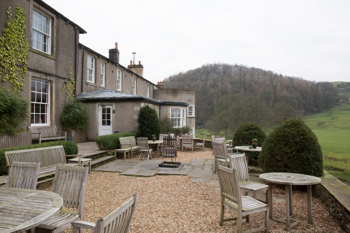 A peaceful outdoor patio at The Inn at Whitewell features wooden tables and chairs arranged on gravel, adjacent to a traditional stone house with ivy-covered walls. The setting overlooks a lush, green landscape in Lancashire, with a hill and trees in the background under a cloudy sky. Image by Andy Wade Photography.