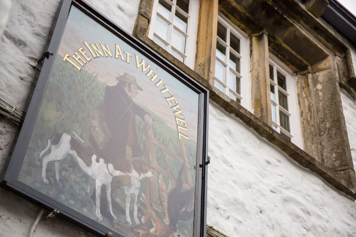 Hanging sign for "The Inn at Whitewell" in Lancashire features a painted depiction of a man in country attire with two dogs, mounted on the white exterior of a building with two windows above. Image by Andy Wade Photography.
