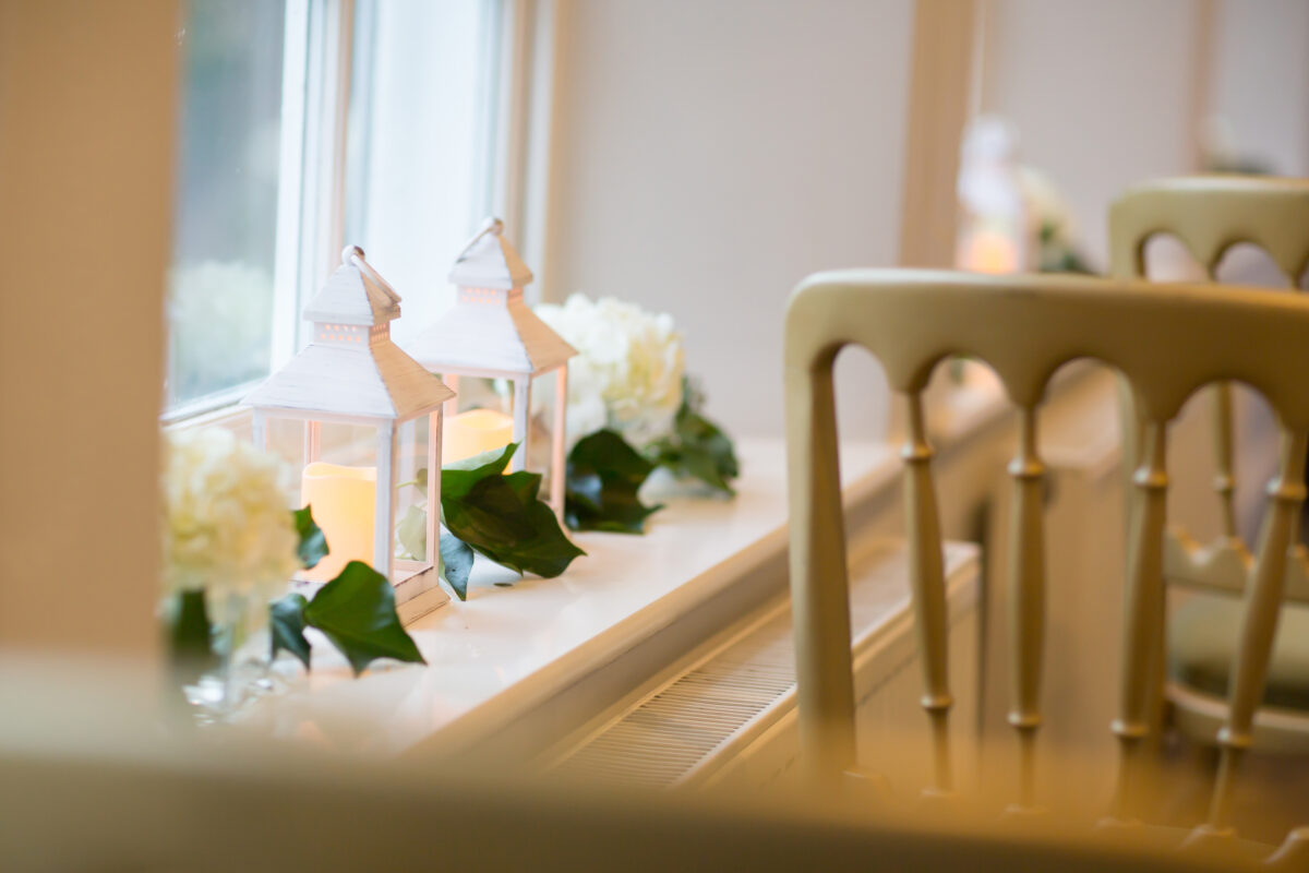 A serene indoor setting at The Inn at Whitewell, Lancashire features two white lanterns with lit candles placed on a windowsill, surrounded by white flowers and green leaves. The soft light from the candles creates a warm and cozy atmosphere. A beige chair is partially visible in the foreground. Image by Andy Wade Photography.