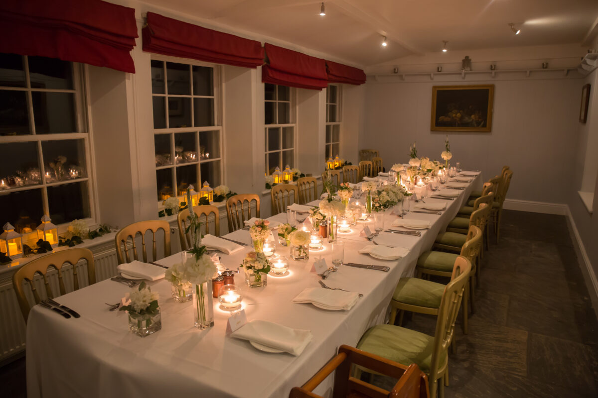 At The Inn at Whitewell in Lancashire, a long dining table is elegantly set up in a dimly lit room for a formal dinner. The table is adorned with white tablecloths, candles, and floral centerpieces. Chairs are placed along both sides, and large windows with red curtains are in the background. Image by Andy Wade Photography.