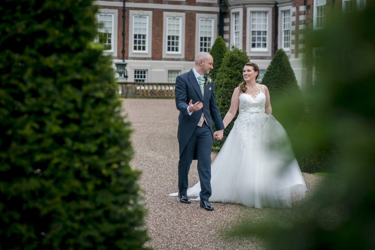 A newlywed couple walks hand in hand outside, dressed in formal wedding attire. The groom is wearing a dark suit and the bride is in a white gown. They are smiling and conversing, surrounded by manicured greenery with Knowsley Hall, Lancashire's stately building, in the background. Image by Andy Wade Photography.
