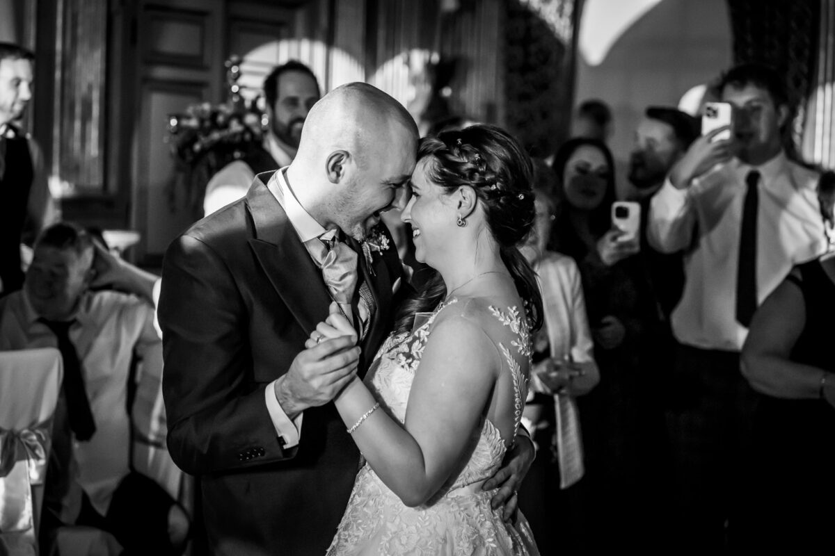A bride and groom share a tender moment during their wedding dance at Knowsley Hall, smiling and touching foreheads. The groom is bald and wearing a suit, while the bride wears a lace gown. Guests in the background smile and take photos. The ornate indoor venue adds a touch of Lancashire charm. Image by Andy Wade Photography.