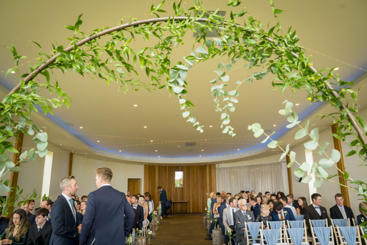 A wedding ceremony setup at The Out Barn in Lancashire with guests seated, facing a pair of grooms standing at the altar. The arch above them is adorned with green foliage. The venue is decorated with string lights and has a modern, elegant design. Image by Andy Wade Photography.