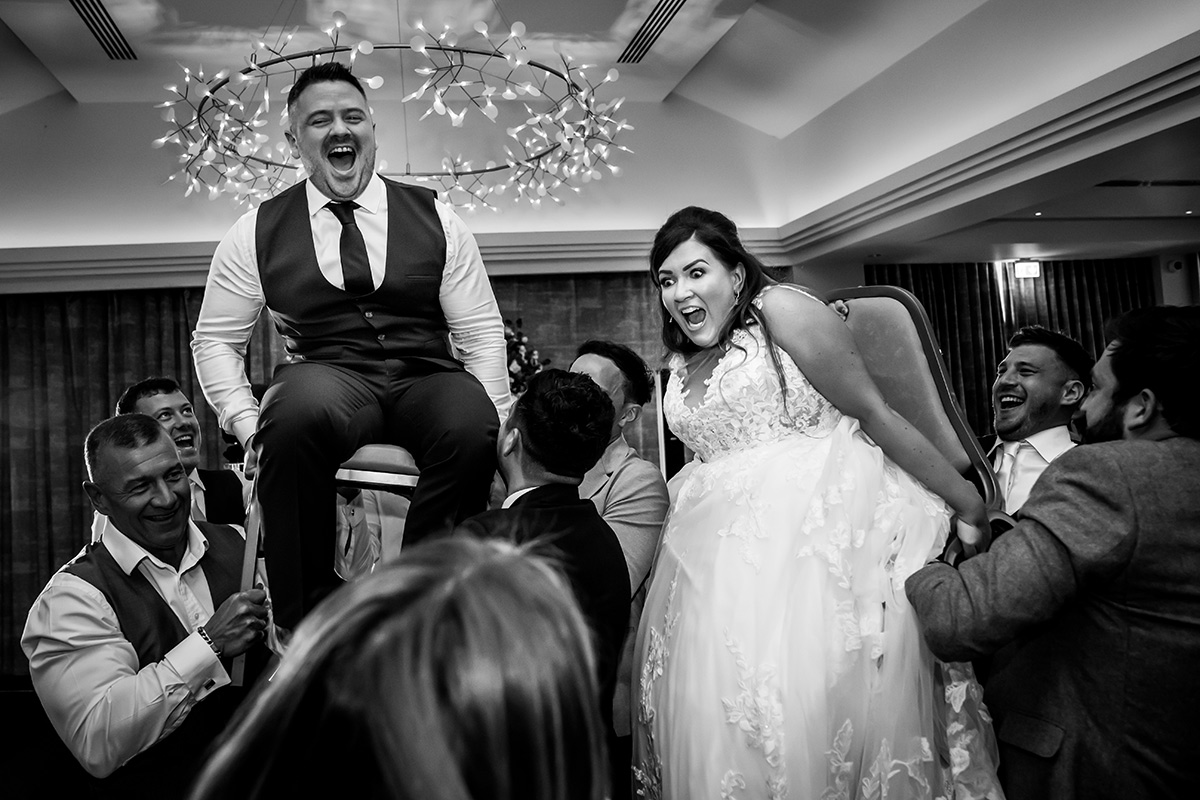 Bride and groom lifted on chairs, guests celebrating.
