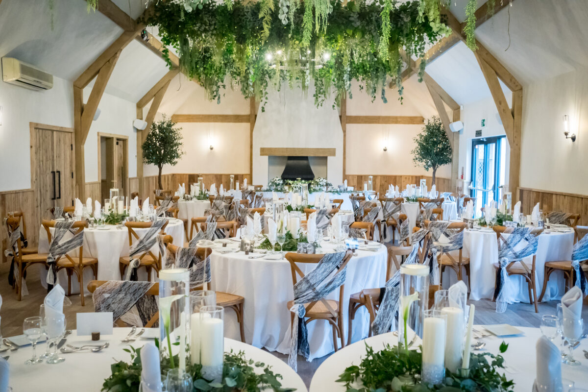 A spacious, elegantly decorated hall at The River Barn in Lancashire set for a wedding reception. Round tables draped with white linens and gray sashes have floral centerpieces and candles. Wooden beams and hanging greenery enhance the ambiance as natural light filters through large windows on the right. Image by Andy Wade Photography.