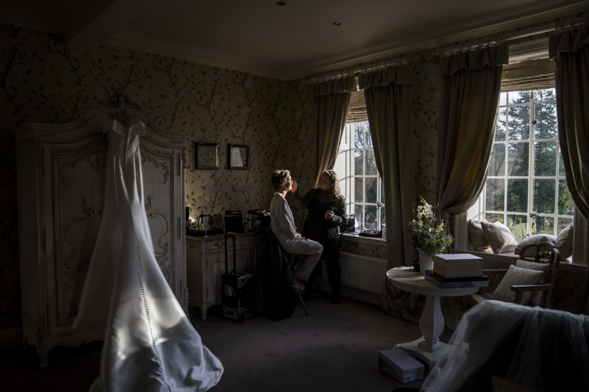 A bride in a white robe sits by a large window with closed curtains in a room adorned with vintage decor at Eaves Hall, Lancashire, receiving makeup from an artist. Her wedding dress hangs elegantly in the foreground. A small table with a plant and some boxes is nearby as sunlight filters gently into the room. Image by Andy Wade Photography.