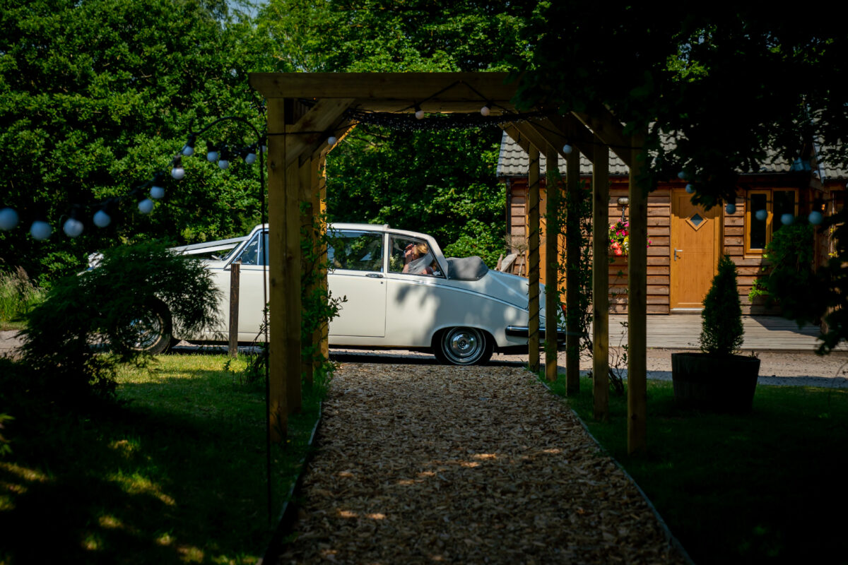A white vintage car is parked on a pathway in front of a wooden cottage at The Tipis at Riley Green, Lancashire, surrounded by lush green foliage. Above the pathway is a wooden canopy adorned with string lights. The scene is set in bright daylight, highlighting the serene and picturesque surroundings. Image by Andy Wade Photography.