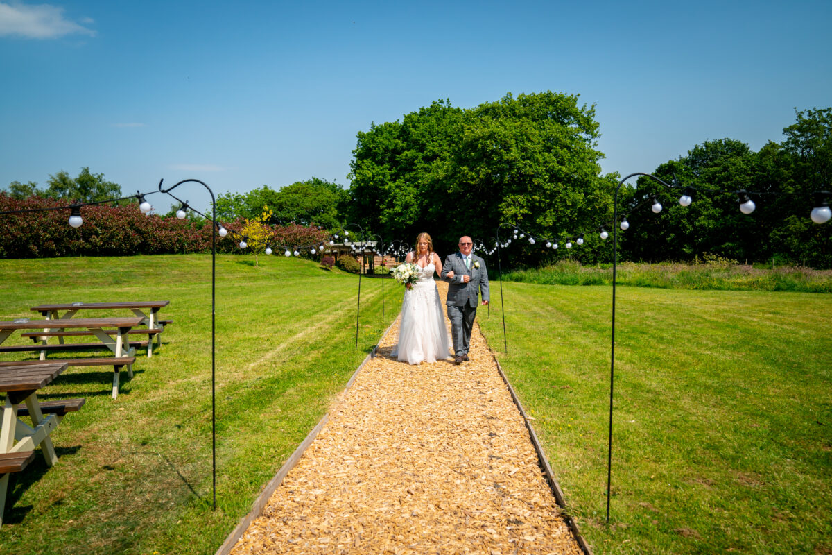 A bride in a white gown walks down an outdoor aisle at The Tipis at Riley Green with an older man in a suit. They are surrounded by a green field, string lights, and picnic tables on a sunny Lancashire day. Image by Andy Wade Photography.