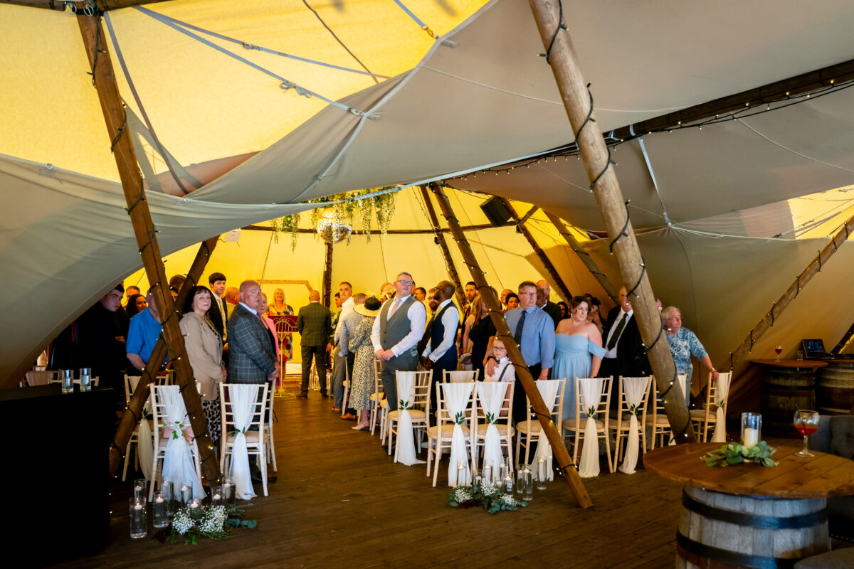 A group of people stand and smile inside a large tent at The Tipis at Riley Green in Lancashire, decorated with greenery and lights. The tent has a wooden frame and white chairs arranged in rows. The atmosphere is cheerful and celebratory. Image by Andy Wade Photography.