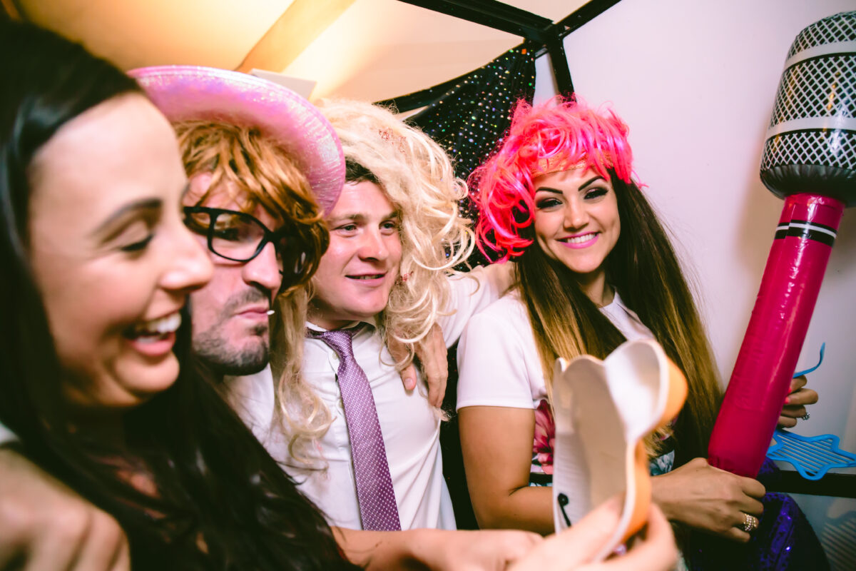 A group of four people dressed in colorful wigs and funny accessories take a selfie together at The White Hart in Lancashire. They are smiling and appear to be having a great time at the event. One holds a large toy microphone while others huddle close, enjoying the moment. Image by Andy Wade Photography.