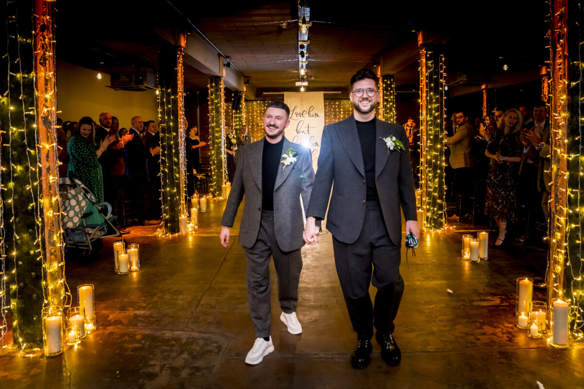 Two grooms walking down candle-lit aisle.
