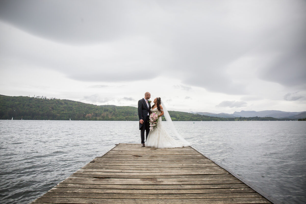 Bride and groom on lakeside pier with clouds.