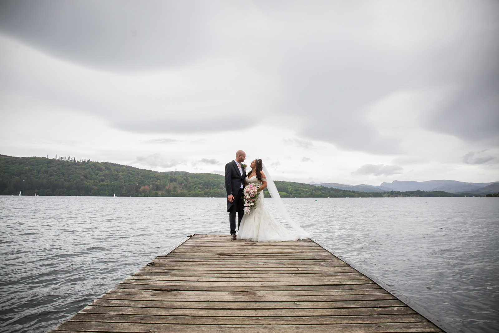 Bride and groom on lakeside pier with clouds.