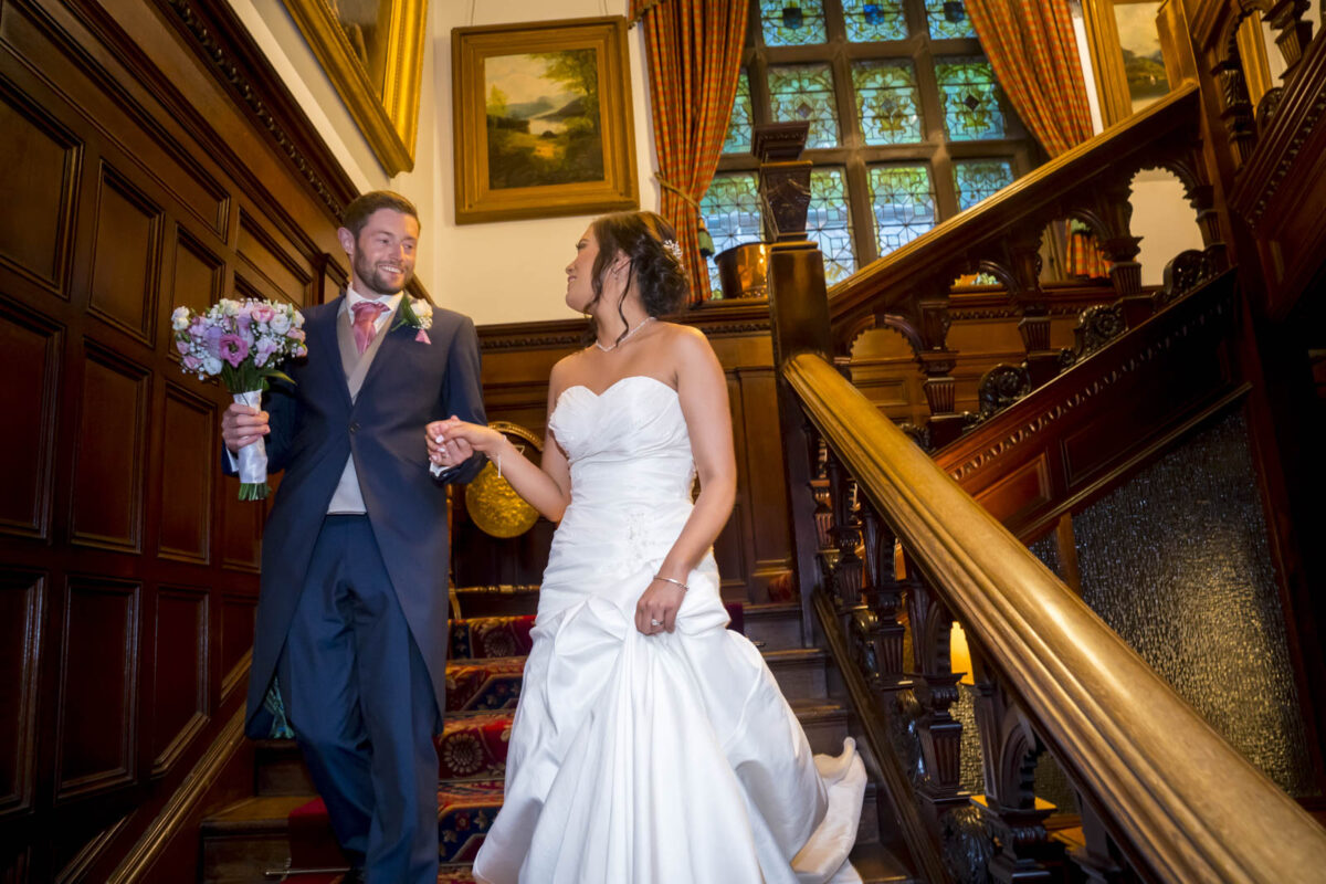 Bride and groom descend ornate wooden staircase.