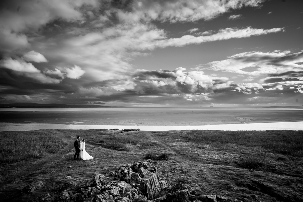 Bride and groom on coastal landscape under dramatic sky.