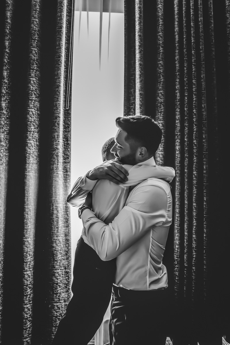 Two people hug near window with curtains.