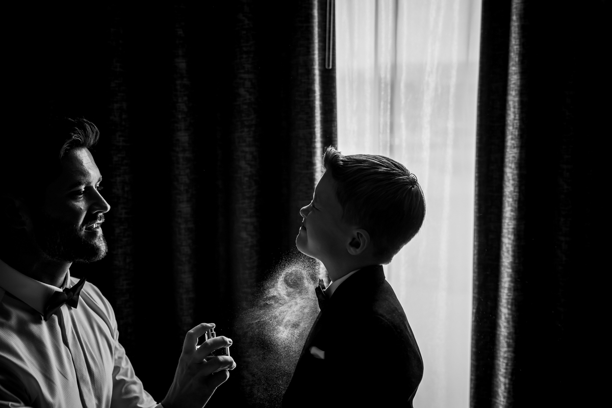 Adult sprays cologne on young boy in suit.