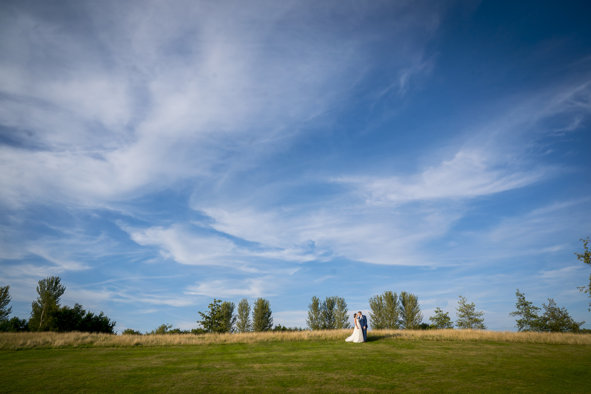 Couple in wedding attire on grassy hill, blue sky.
