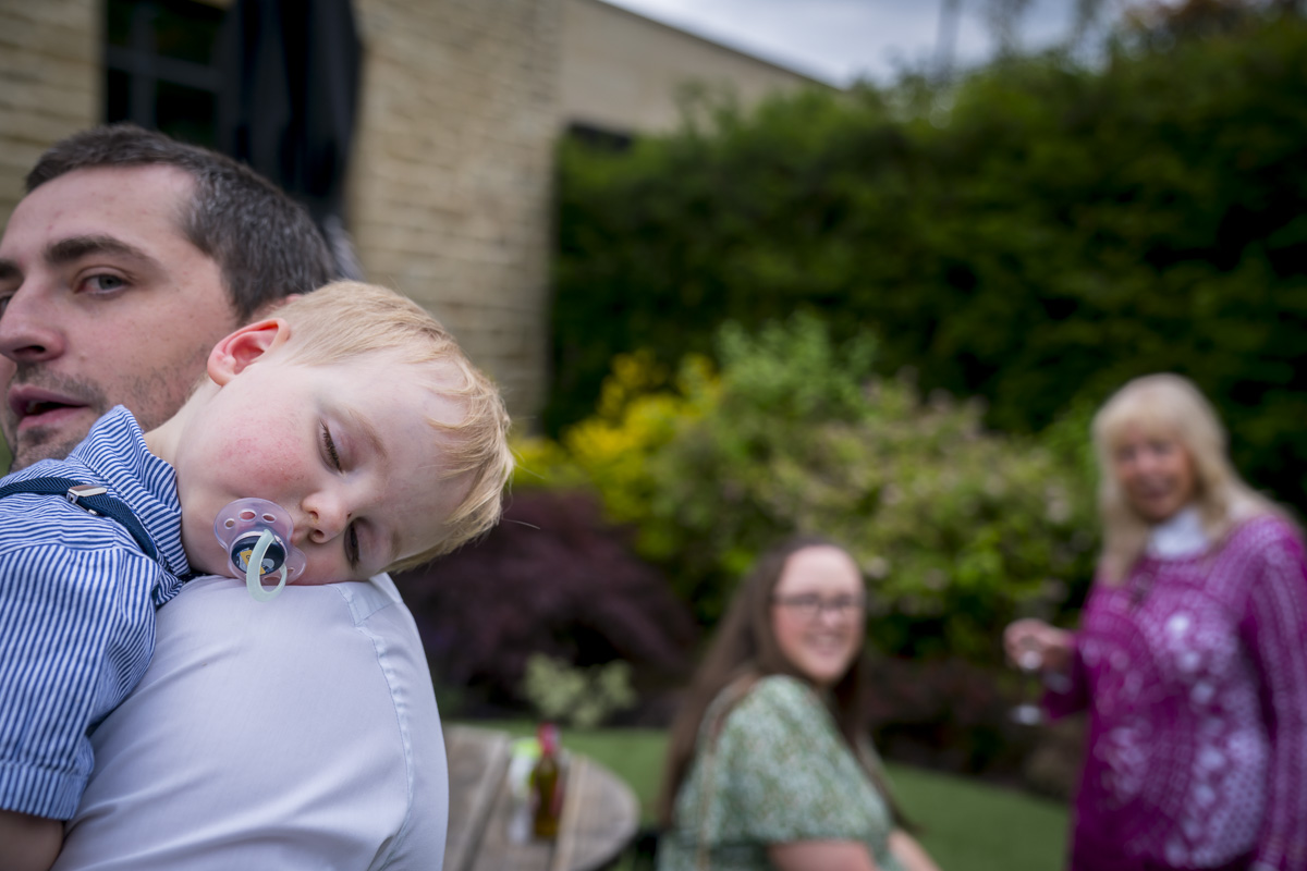 Man holding sleeping child outdoors with people in background.