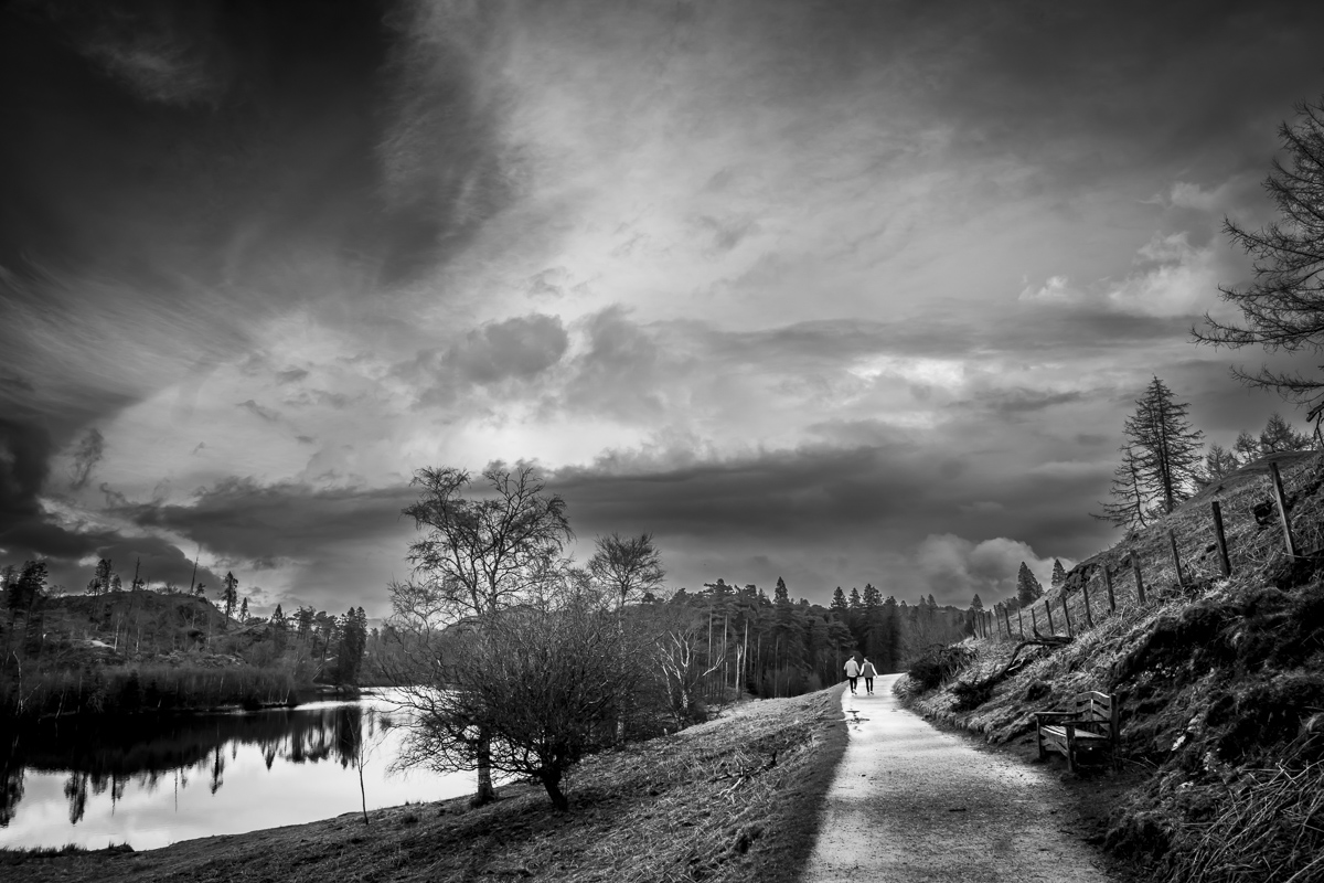 Couple walking along scenic lakeside path under cloudy sky.