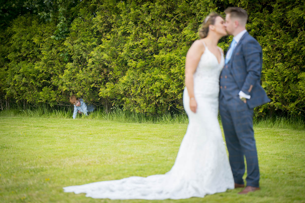 Child peeking from hedge during couple's wedding kiss.