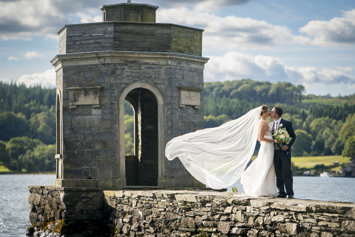 Bride and groom kissing by lake and stone structure.