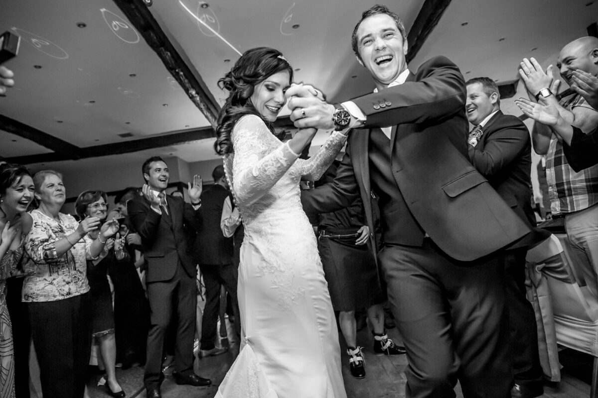 Bride and groom dancing joyfully with guests clapping.
