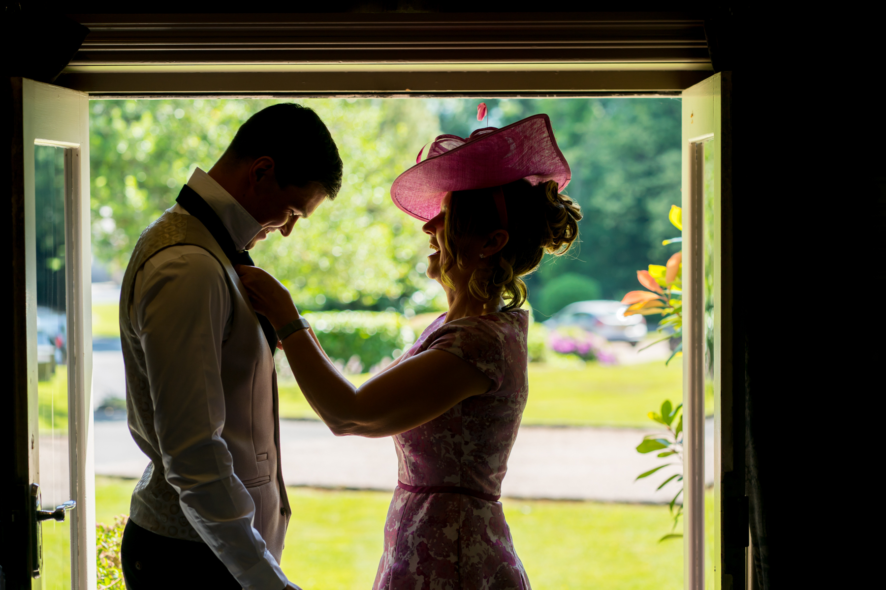 Woman adjusting man's tie at doorway, sunny garden view.