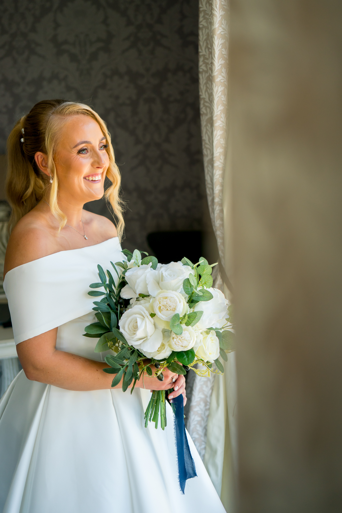 Bride holding white bouquet, smiling by window.