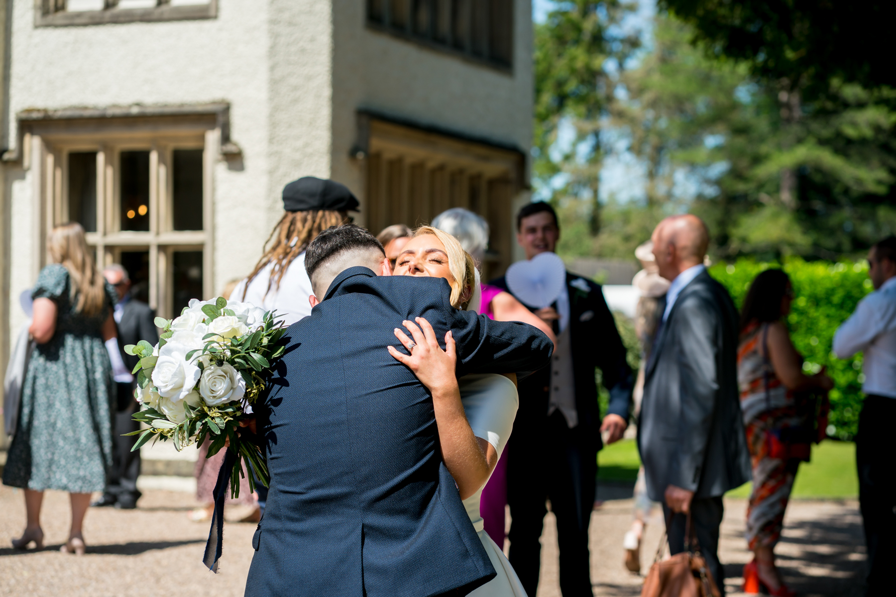 Wedding guests hug outside venue in sunlight.