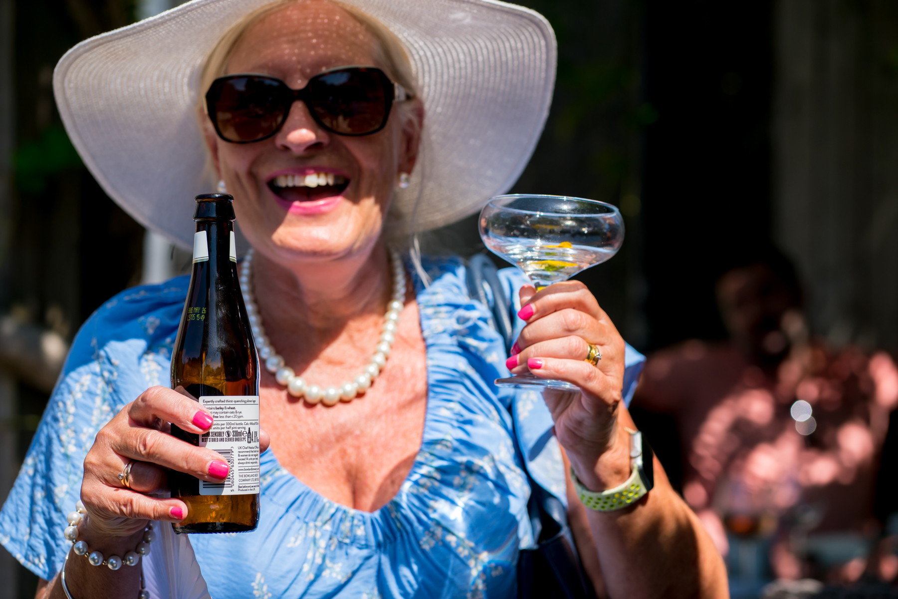 Smiling woman holding drinks outdoors, wearing large hat.