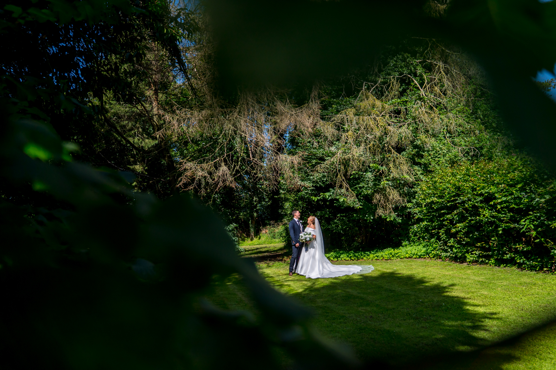 Couple standing in sunlit forest, wedding attire.
