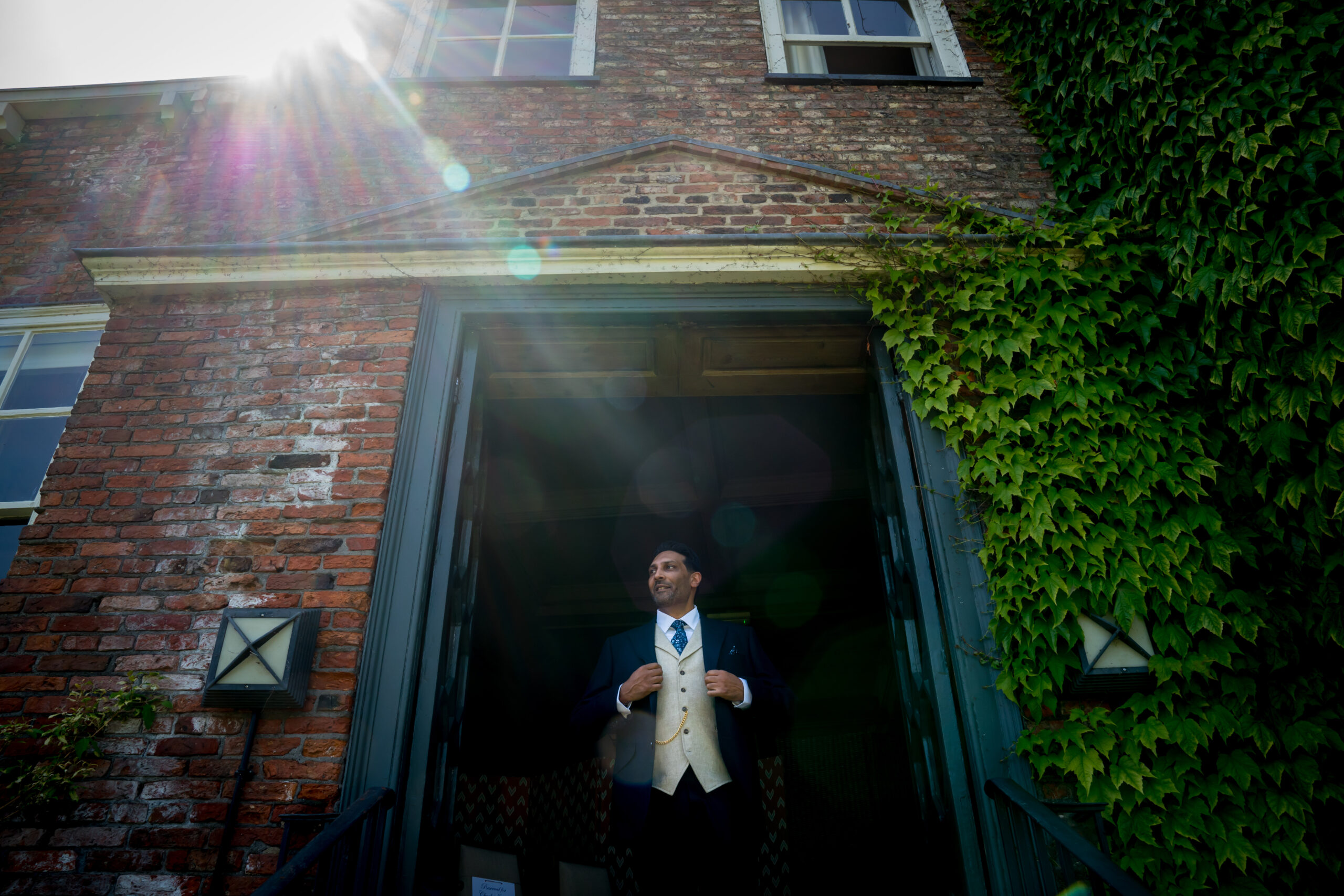 Man in suit standing in sunlight by ivy wall.