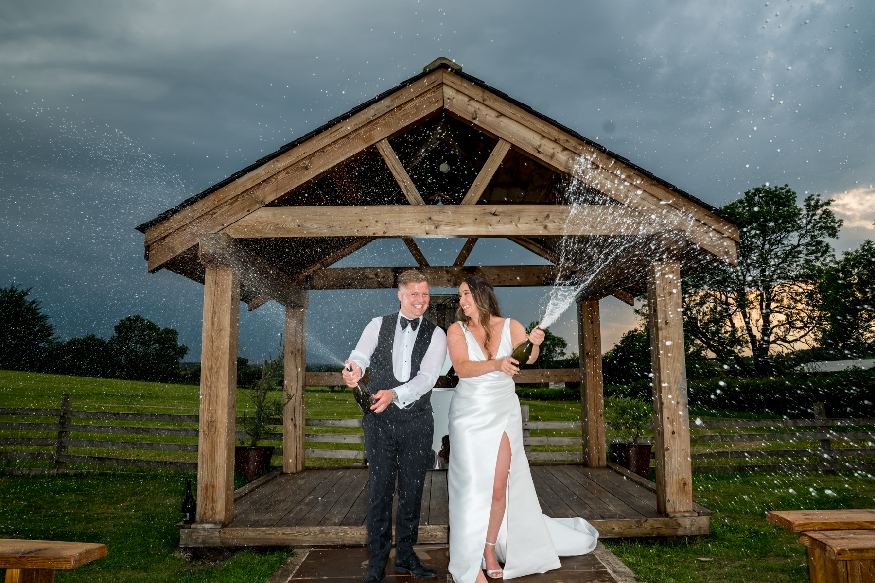 Bride and groom spraying champagne under wooden gazebo